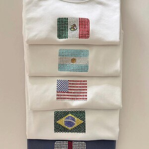 May include: A stack of five folded t-shirts, four white and one dark blue, each featuring a national flag design created with small, sparkling rhinestones. The flags displayed are Mexico, Argentina, the United States, Brazil, and England.