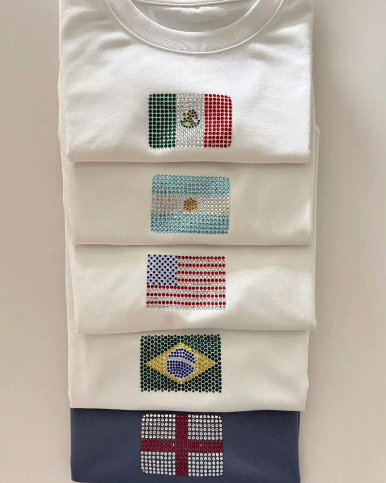 May include: A stack of five folded t-shirts, four white and one dark blue, each featuring a national flag design created with small, sparkling rhinestones. The flags displayed are Mexico, Argentina, the United States, Brazil, and England.