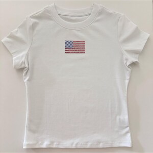 May include: A white short-sleeved t-shirt featuring a small, centered graphic of the United States flag. The flag design is composed of individual red, white, and blue rhinestones, creating a textured, sparkling appearance on the plain fabric.