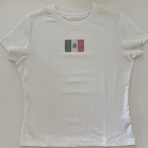 May include: A white short-sleeved t-shirt featuring a small, centered rectangular graphic of the Mexican flag. The flag design is composed of individual green, white, and red rhinestones, with a central emblem detail in the white section.