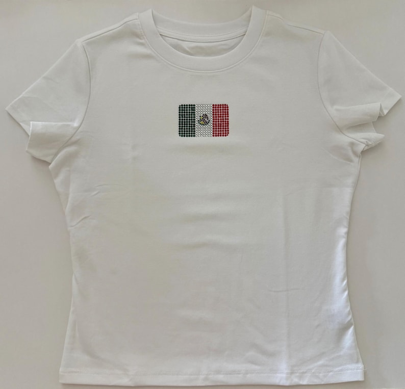 May include: A white short-sleeved t-shirt featuring a small, centered rectangular graphic of the Mexican flag. The flag design is composed of individual green, white, and red rhinestones, with a central emblem detail in the white section.