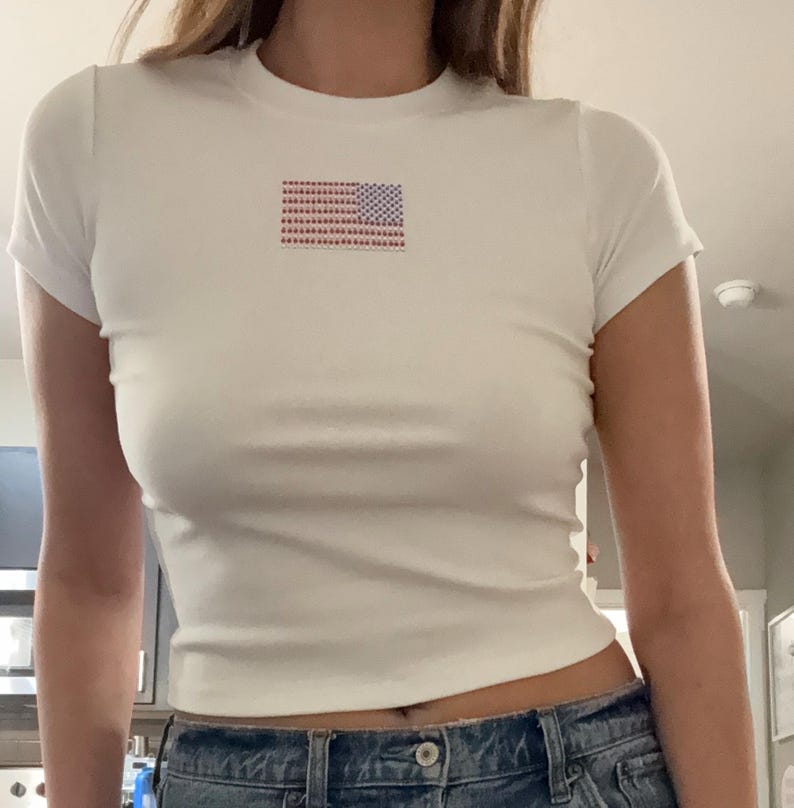May include: A white short-sleeved t-shirt featuring a small, centered graphic of the American flag composed of tiny red, white, and blue rhinestones. The shirt has a crew neckline and a fitted silhouette, paired with blue denim jeans.