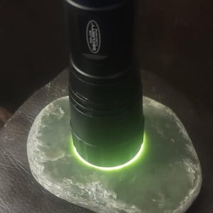 Può includere: Una torcia nera con un accento blu illumina una pietra ovale verde chiaro. Una moneta è accanto alla pietra. La torcia ha le parole "POLICE SECURITY".