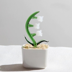 May include: A glass lily of the valley plant in a white square ceramic pot. The plant features a green stem and leaves with three white bell-shaped flowers. The pot is filled with small, light-colored pebbles.