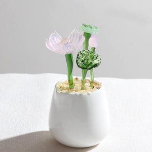 May include: A white ceramic pot with glass flowers. The flowers are pink, green, and clear, with green stems. The pot is filled with small, light-colored pebbles.