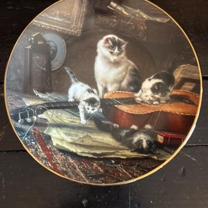 May include: Decorative plate featuring a detailed painting of four cats near a guitar. The cats are white and gray, with one lying on its back. The plate has a gold rim and depicts a still life scene with musical instruments.