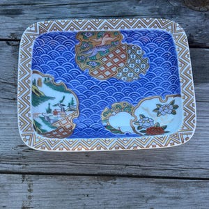 May include: A rectangular ceramic tray featuring a blue wave pattern background with three distinct decorative panels. The panels display a landscape with trees and buildings, a flying bird, and floral motifs, all bordered by a gold geometric pattern.