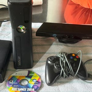 以下が含まれることがあります： 黒いXbox 360本体、電源アダプター、黒いKinectセンサーバー、色付きボタンを備えた有線の黒いゲームコントローラー、Just Dance 2014のゲームディスク。本体には緑色に光る電源ボタンとXbox 360のロゴが付いています。