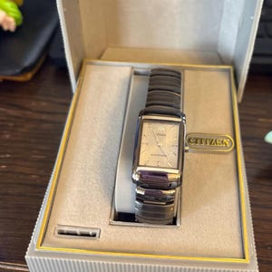 May include: A Citizen watch in its original box. The watch has a rectangular silver face with black bands. The watch is in a gray box with the Citizen logo in gold lettering.