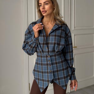 May include: A blue plaid shirt with a unique design. The shirt features a button-down front, long sleeves, and a cinched waist. The plaid pattern includes shades of blue, brown, and white. The shirt is paired with brown leggings.