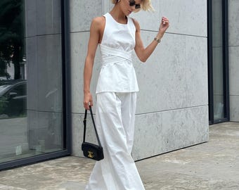 Puede incluir: Un conjunto blanco compuesto por un top sin mangas con diseño cruzado y pantalones blancos de pernera ancha. El conjunto se complementa con un pequeño bolso negro y sandalias negras. La persona lleva gafas de sol y un reloj.
