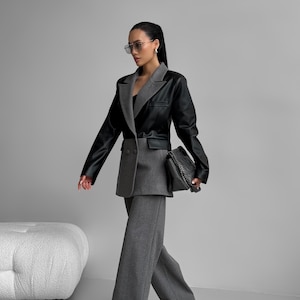 Könnte beinhalten: Ein stilvolles Outfit aus schwarzem und grauem Anzug. Die Jacke hat einen oberen Teil aus schwarzem Leder und einen unteren Teil aus grauem Wollstoff, kombiniert mit passenden Hosen mit weitem Bein. Das Outfit wird durch schwarze spitze Absätze und eine Handtasche mit Kette abgerundet. Der Look ist modern und schick.