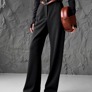 May include: A person wearing a black long-sleeve top and black wide-leg pants. A brown leather belt with a silver buckle is around the waist. A brown, circular handbag with a zipper is held in the hand. Black pointed-toe shoes complete the outfit.
