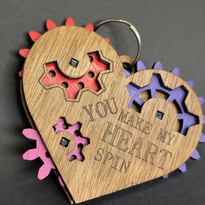 May include: Wooden heart-shaped keychain with the engraved text "YOU MAKE MY HEART SPIN." The keychain features a gear design with red, purple, and pink accents. A silver key ring is attached to the top.