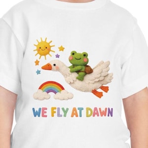 May include: White t-shirt with a colourful illustration. A green frog is riding a white goose, with a rainbow, sun, and stars. The text "WE FLY AT DAWN" is printed in various colours. A fun and playful design for children.