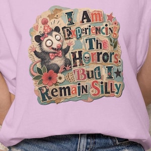 May include: A light purple t-shirt featuring a graphic of a wide-eyed opossum wearing a party hat and bowtie. The text reads, I Am Experiencing The Horrors, But I Remain Silly. The design includes floral accents, a small skull, and stars.