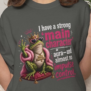 Könnte beinhalten: Ein anthrazitfarbenes T-Shirt mit einer Grafik eines grünen Frosches, der eine goldene Krone, eine rosa herzförmige Sonnenbrille und eine rosa Pelzstola trägt. Der Frosch sitzt auf einem roten Kissen mit dem Text: I have a strong 'main character' aura—and almost no impulse control.