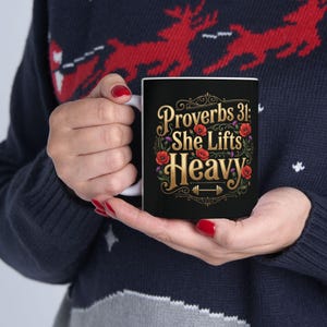 Könnte beinhalten: Schwarze Keramik-Tasse mit dem Text "Proverbs 31: She Lifts Heavy" in goldfarbenen Buchstaben, akzentuiert mit roten floralen Designs. Die Tasse hat einen weißen Griff und eine Hantel-Grafik. Die Tasse wird von einer Person gehalten, die einen dunkelblauen Pullover trägt.