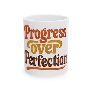 Könnte beinhalten: Eine weiße Keramiktasse mit dem Schriftzug "Progress Over Perfection" in einem Retro-Schriftstil. Der Text ist in Orange- und Brauntönen gehalten. Die Tasse hat eine Standardgröße und -form.