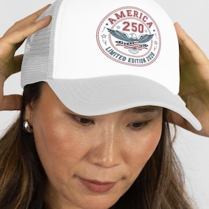 Könnte beinhalten: Eine weiße Trucker-Cap mit einer kreisförmigen Grafik auf der Vorderseite. Das Design zeigt einen Weißkopfseeadler mit US-Flaggenmuster sowie den Text AMERICA 250 und LIMITED EDITION 2026, eingefasst in einen roten, seilartigen Rand.