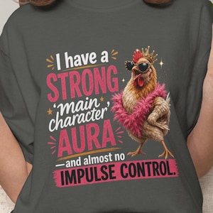 Könnte beinhalten: Ein dunkelgraues T-Shirt mit einer Grafik eines Huhns, das eine goldene Krone, eine schwarze Sonnenbrille und eine rosa Federboa trägt. Der Text lautet: I have a STRONG main character AURA and almost no IMPULSE CONTROL in weißer und rosa Schrift.