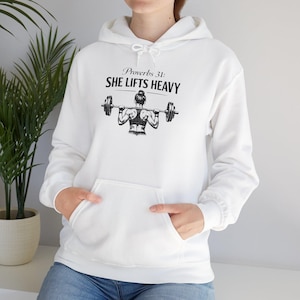 Könnte beinhalten: Weißer Kapuzenpullover mit Fronttasche und Kordelzug. Der Pullover zeigt den Text "Proverbs 31: SHE LIFTS HEAVY" über einer Grafik eines Gewichthebers, der eine Langhantel hebt. Das Design ist in Schwarz.