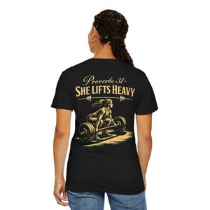 Könnte beinhalten: Schwarzes T-Shirt mit goldfarbener Grafik einer Frau, die eine Langhantel hebt. Der Text "Proverbs 31- SHE LIFTS HEAVY" befindet sich über der Illustration. Das Shirt besteht aus weichem Material und ist für Workouts oder Freizeit geeignet.