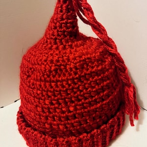 May include: A handmade, red crochet hat with a rounded crown and a ribbed brim. A long, twisted cord extends from the top of the hat. The hat is made of a thick yarn, and the stitches are clearly visible. The hat is a solid, vibrant red color.