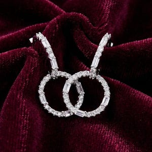 Peut inclure: Une paire de boucles d'oreilles créoles argentées avec un design étincelant orné de pierres précieuses. Les boucles d'oreilles présentent un cercle avec une section plus petite ornée de pierres précieuses rectangulaires. Présentées sur du velours rouge foncé.
