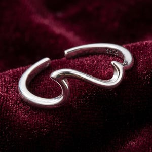 Può includere: Un anello aperto in argento con un design liscio a forma di onda. La fascia in metallo ha una finitura lucida e include il marchio S925 impresso chiaramente sulla superficie interna, indicando la composizione del gioiello.