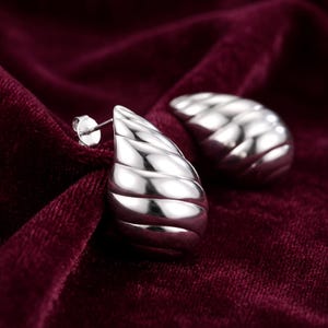 May include: A pair of polished silver-tone stud earrings featuring a teardrop shape with a series of horizontal, curved ridges that create a shell-like texture. Each earring includes a standard post back mechanism for secure attachment to the earlobe.
