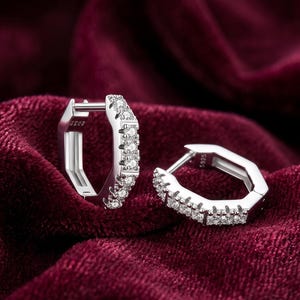 May include: A pair of silver-toned hoop earrings with a geometric design. Each earring is encrusted with small, sparkling gemstones along one side. The earrings are displayed on a deep red velvet surface.