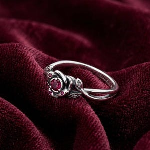May include: A silver ring featuring a rose design with a central red gemstone and smaller red accents. The ring has a delicate leaf detail and is set against a deep red velvet background. The brand name "PANDORA" is visible on the ring.