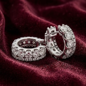May include: A pair of silver-toned hoop earrings featuring two parallel rows of round, clear, faceted stones set into the metal. The earrings have a hinged closure mechanism and a polished finish, showcasing a circular design with multiple stones.