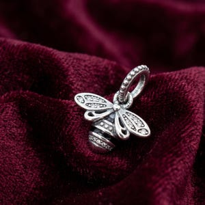 Pode incluir: Um berloque de abelha prateada com detalhes de cristal transparente nas asas e no corpo. O berloque tem um design detalhado e uma argola para prender a uma pulseira ou colar. O abdómen da abelha apresenta riscas de esmalte preto.
