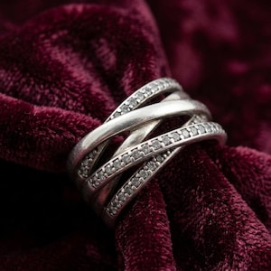 Puede incluir: Un anillo plateado con un diseño entrelazado, engastado con pequeñas piedras brillantes. El anillo se muestra sobre una superficie de terciopelo rojo oscuro, creando un contraste.