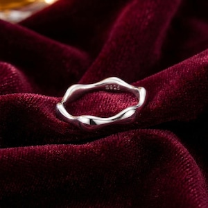 May include: A silver ring with a flowing, irregular shape. The ring is crafted from a bright metal and has a smooth, reflective finish. The inside of the ring is marked with "S925". It rests on a deep red velvet surface.