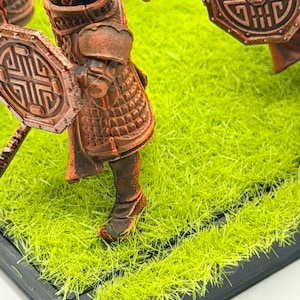 K&ouml;nnte beinhalten: Miniaturfigur eines Kriegers in bronzefarbener R&uuml;stung, der einen Schild mit geometrischem Design h&auml;lt. Die Figur steht auf einer Basis aus k&uuml;nstlichem, gr&uuml;nem Gras. Die R&uuml;stung hat detaillierte Muster.