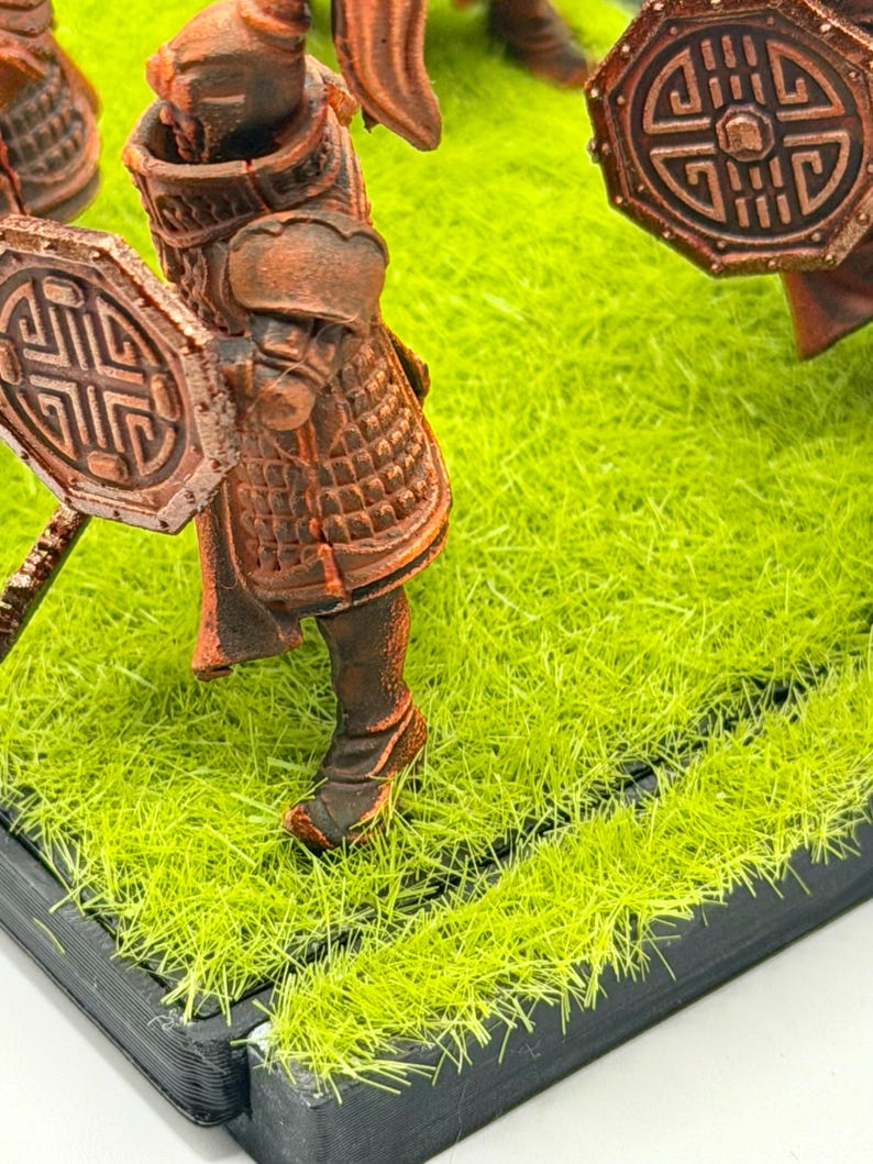Może przedstawiać: Miniaturowa figurka wojownika w brązowej zbroi, trzymającego tarczę z geometrycznym wzorem. Figurka stoi na podstawie ze sztucznej zielonej trawy. Zbroja ma skomplikowane detale.