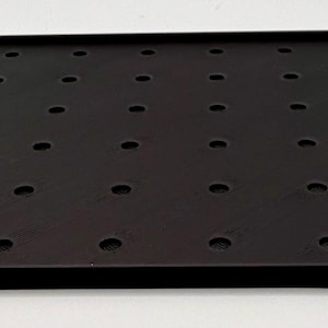 K&ouml;nnte beinhalten: Ein schwarzes quadratisches Tablett mit erh&ouml;htem Rand und zahlreichen, gleichm&auml;&szlig;ig verteilten kreisf&ouml;rmigen L&ouml;chern. Das Tablett besteht aus einem matten Material und liegt auf einer wei&szlig;en Oberfl&auml;che. Die Abmessungen des Tabletts betragen ca. 30 cm x 30 cm.
