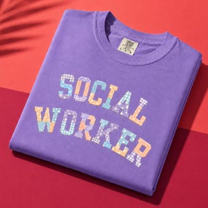 以下が含まれることがあります： パッチワークデザインで「SOCIAL WORKER」の文字が入った紫色のTシャツ。テキストは様々なパステルカラーとパターンで表現されています。折りたたまれたTシャツは、赤とオレンジの表面に置かれています。