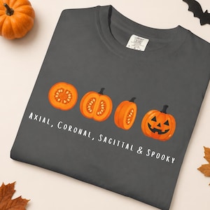 May include: A charcoal gray t-shirt featuring a graphic of four orange pumpkins in various cross-sections and a jack-o'-lantern. The text printed below the pumpkins reads: Axial, Coronal, Sagittal & Spooky. The shirt has a crew neckline.