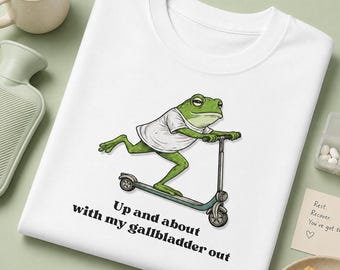 May include: A white t-shirt featuring a green frog wearing a white t-shirt and riding a grey scooter. Below the illustration, the black text reads: Up and about with my gallbladder out. The shirt is displayed on a flat surface with a mug and crackers.