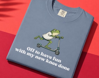 May include: A blue t-shirt features a cartoon frog riding a scooter. The frog wears a white shirt and shorts. Text on the shirt reads "Off to have fun with my new knee done."
