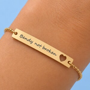 May include: A gold-colored bracelet with the inscription "Bendy not broken" and a heart-shaped cutout. The bracelet is on a wrist against a light blue background. The bracelet is made of a delicate chain.