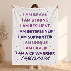 May include: A white blanket with the phrases "I AM BRAVE", "I AM STRONG", "I AM RESILIENT", "I AM DETERMINED", "I AM SUPPORTED", "I AM UNIQUE", "I AM LOVED", "I AM A CF WARRIOR", and "I AM OLIVIA" in purple and blue text. The blanket has small, colorful polka dots.