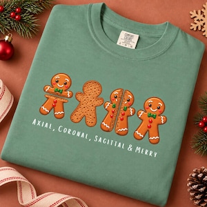 May include: A sage green t-shirt featuring a graphic of four gingerbread cookies in various states of anatomical sectioning, with the text "Axial, Coronal, Sagittal & Merry" printed in white below the cookies. The shirt has a crew neck collar.