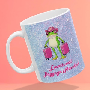 May include: White ceramic mug with a cartoon frog in a pink cowboy hat, holding two pink suitcases. The mug has a light blue glitter background and the text "Emotional Baggage Handler" in pink script.