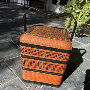 May include: A vintage rectangular picnic basket with a woven rattan design in shades of orange and black. The basket has a hinged lid and a black metal handle for carrying. The basket is sitting on a stone surface.