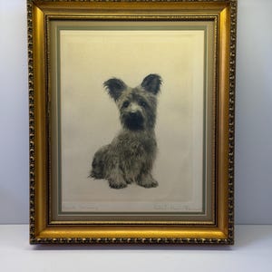 May include: A framed print of a seated dog, rendered in shades of gray and black. The dog has long, floppy ears and a fluffy coat. The frame is ornate, with a gold finish and decorative detailing. The print is signed.